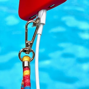 Puede incluir: Una funda de tel&eacute;fono roja con un cable de carga blanco conectado a un broche dorado y un cord&oacute;n de rayas de colores.
