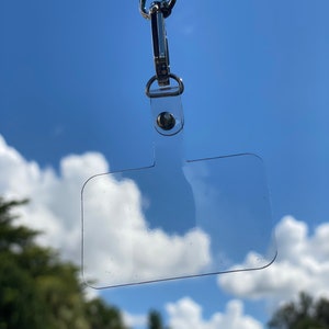 May include: A clear plastic ID badge holder with a silver clip and a blue lanyard. The holder is hanging from the lanyard against a blue sky with white clouds.