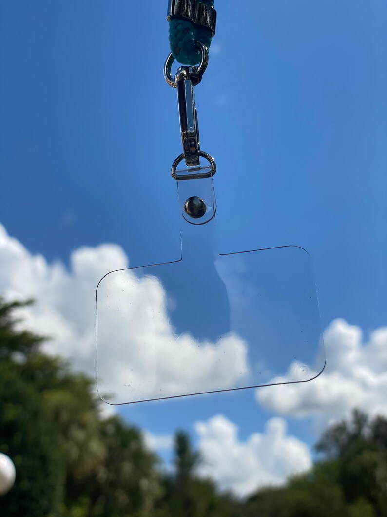 May include: A clear plastic ID badge holder with a silver clip and a blue lanyard. The holder is hanging from the lanyard against a blue sky with white clouds.