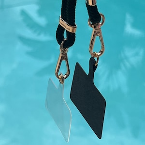 May include: Two black straps with gold clasps and a gold ring with the word "ATHOMElace" engraved on it. The straps are attached to two rectangular tags, one black and one clear.