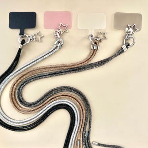 Może przedstawiać: Cztery różne smycze do telefonu z zawieszkami w kształcie gwiazd. Każda smycz wykonana jest z błyszczącego sznurka w kolorze czarnym, srebrnym, różowym złocie i złotym. Smycze są prezentowane na beżowym tle.