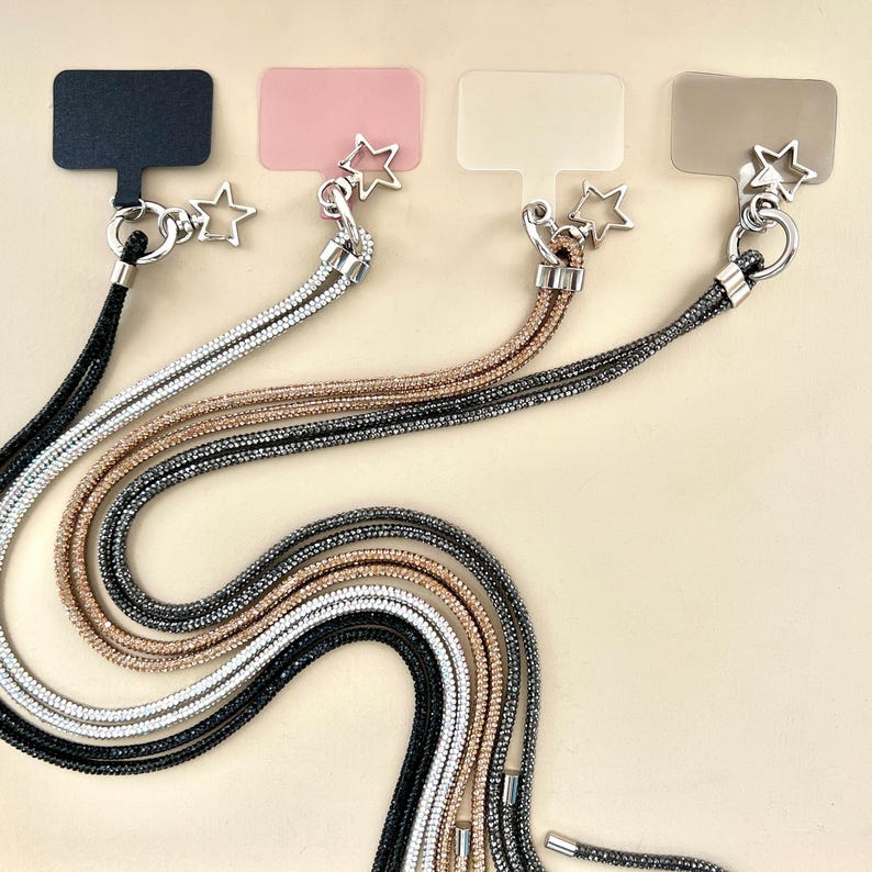 May include: Four different phone straps with star charms. Each strap is made of a sparkly cord in black, silver, rose gold, and gold. The straps are displayed on a beige background.