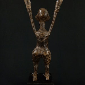 K&ouml;nnte beinhalten: Bronzestatue einer Figur mit erhobenen Armen, die auf einem schwarzen Sockel steht. Die Figur hat detaillierte Schnitzereien am Torso und an den Armen. Die Statue steht vor einem dunklen Hintergrund.