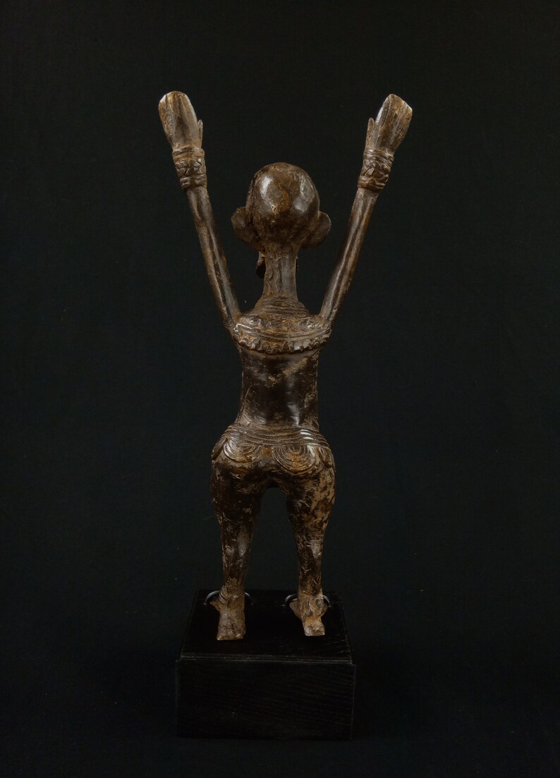 K&ouml;nnte beinhalten: Bronzestatue einer Figur mit erhobenen Armen, die auf einem schwarzen Sockel steht. Die Figur hat detaillierte Schnitzereien am Torso und an den Armen. Die Statue steht vor einem dunklen Hintergrund.