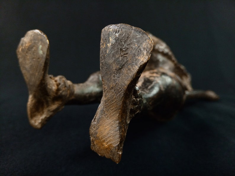 K&ouml;nnte beinhalten: Eine Bronzeskulptur einer liegenden Figur, von unten betrachtet. Die F&uuml;&szlig;e der Figur sind deutlich sichtbar, mit strukturierten Oberfl&auml;chen und einer dunklen, gealterten Patina. Die Skulptur ist vor einem dunklen Hintergrund platziert.