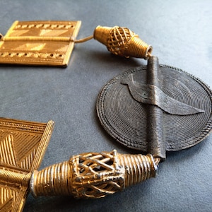 Peut inclure: Un collier en or et noir avec un pendentif circulaire représentant un oiseau stylisé. Le collier est fait de fil métallique et a une texture tissée.