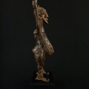 K&ouml;nnte beinhalten: Eine detaillierte, braune Holzskulptur einer Figur mit erhobenen Armen. Die Figur hat einen Bart und aufwendige Schnitzereien auf Brust und Beinen. Sie steht auf einem schwarzen Sockel vor dunklem Hintergrund. Wahrscheinlich afrikanische Stammeskunst.