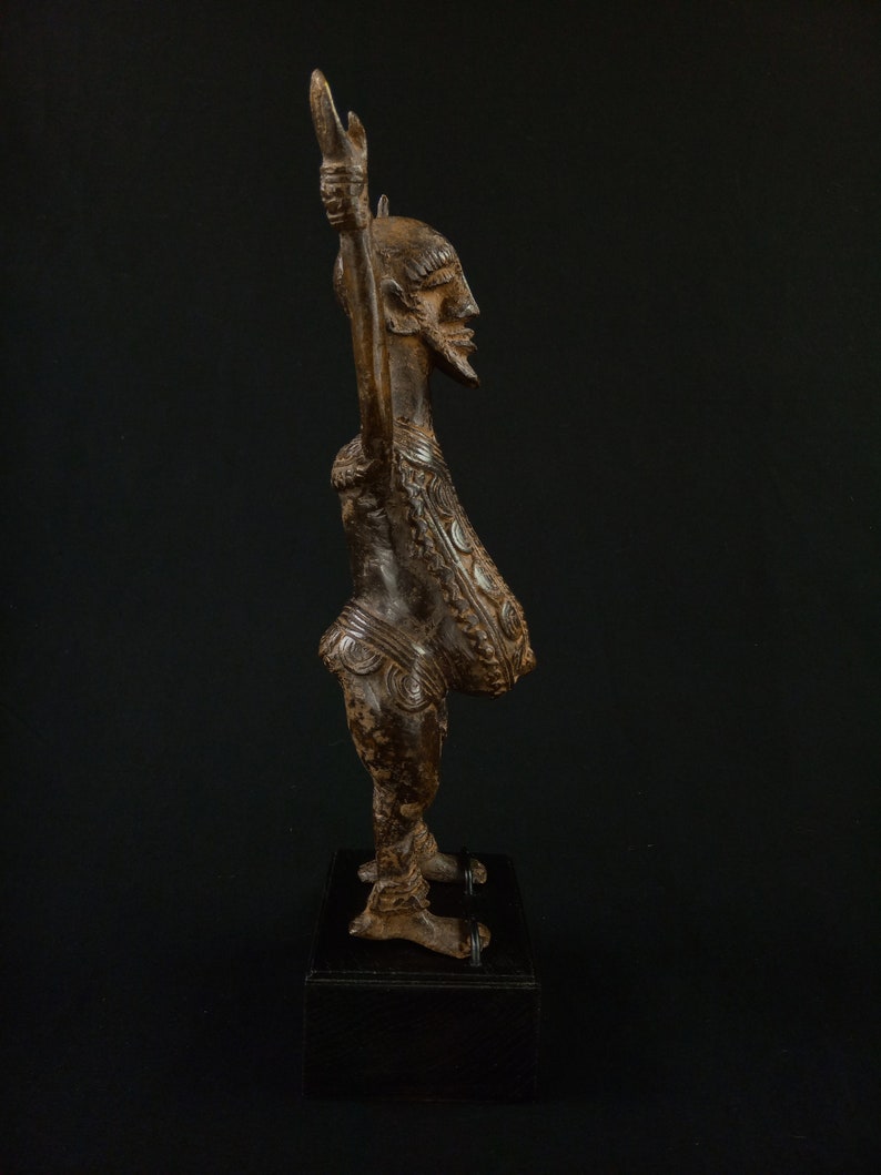 K&ouml;nnte beinhalten: Eine detaillierte, braune Holzskulptur einer Figur mit erhobenen Armen. Die Figur hat einen Bart und aufwendige Schnitzereien auf Brust und Beinen. Sie steht auf einem schwarzen Sockel vor dunklem Hintergrund. Wahrscheinlich afrikanische Stammeskunst.