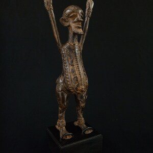 K&ouml;nnte beinhalten: Braune Holzstatue einer Figur mit erhobenen Armen. Die Figur hat ein detailliertes Gesicht und einen detaillierten K&ouml;rper und steht auf einem schwarzen Sockel. Die Statue ist wahrscheinlich ein dekoratives Kunstwerk.