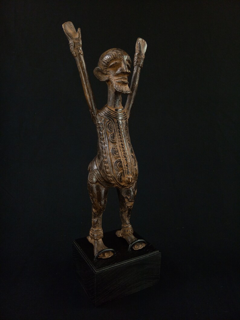 K&ouml;nnte beinhalten: Braune Holzstatue einer Figur mit erhobenen Armen. Die Figur hat ein detailliertes Gesicht und einen detaillierten K&ouml;rper und steht auf einem schwarzen Sockel. Die Statue ist wahrscheinlich ein dekoratives Kunstwerk.