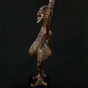 K&ouml;nnte beinhalten: Eine detaillierte Bronzeskulptur einer Figur mit langem Hals und erhobenem Arm. Die Figur hat einen Bart und komplizierte Muster auf dem Torso. Sie steht auf einem dunklen Holzsockel vor schwarzem Hintergrund.