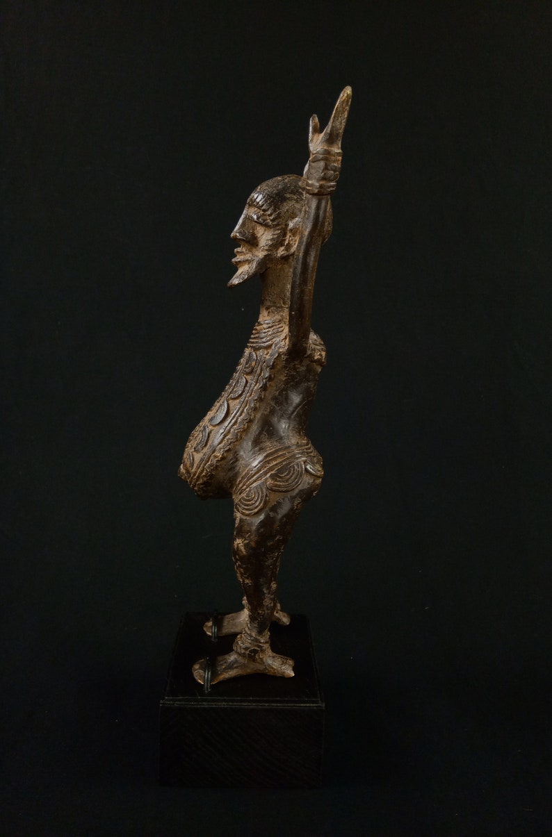 K&ouml;nnte beinhalten: Eine detaillierte Bronzeskulptur einer Figur mit langem Hals und erhobenem Arm. Die Figur hat einen Bart und komplizierte Muster auf dem Torso. Sie steht auf einem dunklen Holzsockel vor schwarzem Hintergrund.