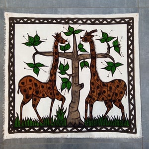May include: A white canvas with a black border depicting two brown giraffes with black spots standing near a green tree with brown leaves. The giraffes are facing each other and the tree is in the center of the canvas.