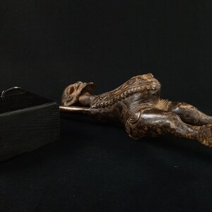 K&ouml;nnte beinhalten: Eine dunkelbraune, geschnitzte Holzfigur einer liegenden Person mit aufwendigen Details. Die Figur liegt auf einer Metallstange, im Hintergrund eine schwarze Box mit zwei Metallgriffen. Die Figur befindet sich auf einer schwarzen Oberfl&auml;che.