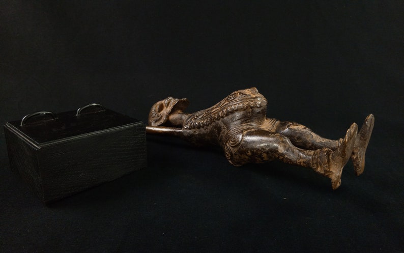 K&ouml;nnte beinhalten: Eine dunkelbraune, geschnitzte Holzfigur einer liegenden Person mit aufwendigen Details. Die Figur liegt auf einer Metallstange, im Hintergrund eine schwarze Box mit zwei Metallgriffen. Die Figur befindet sich auf einer schwarzen Oberfl&auml;che.