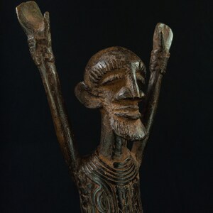 K&ouml;nnte beinhalten: Eine detaillierte, dunkelbraune Skulptur einer Figur mit erhobenen Armen. Die Figur hat einen Bart und detaillierte Gesichtsz&uuml;ge. Die Kleidung der Skulptur ist mit komplizierten Mustern verziert. Der Hintergrund ist schwarz.