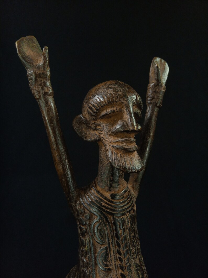 K&ouml;nnte beinhalten: Eine detaillierte, dunkelbraune Skulptur einer Figur mit erhobenen Armen. Die Figur hat einen Bart und detaillierte Gesichtsz&uuml;ge. Die Kleidung der Skulptur ist mit komplizierten Mustern verziert. Der Hintergrund ist schwarz.