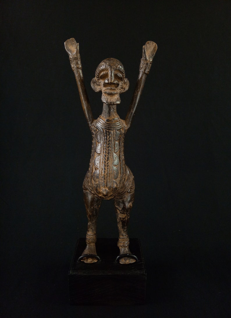 K&ouml;nnte beinhalten: Bronzefarbene Skulptur einer menschlichen Figur mit erhobenen Armen. Die Figur hat ein detailliertes Gesicht und einen Torso mit gemusterten Designs. Sie steht auf einem schwarzen rechteckigen Sockel.