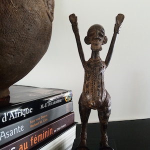 K&ouml;nnte beinhalten: Eine bronzefarbene Skulptur einer stilisierten menschlichen Figur mit erhobenen Armen. Die Figur steht auf einem schwarzen Sockel, mit detaillierten Mustern am Torso. B&uuml;cher mit franz&ouml;sischen Titeln sind in der N&auml;he gestapelt, und ein gro&szlig;es, strukturiertes, dunkelbraunes Gef&auml;&szlig; ist teilweise sichtbar.