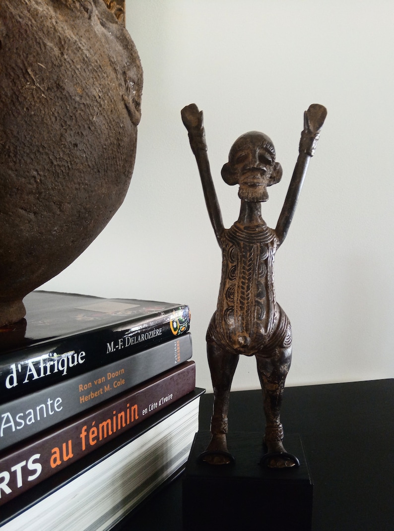 K&ouml;nnte beinhalten: Eine bronzefarbene Skulptur einer stilisierten menschlichen Figur mit erhobenen Armen. Die Figur steht auf einem schwarzen Sockel, mit detaillierten Mustern am Torso. B&uuml;cher mit franz&ouml;sischen Titeln sind in der N&auml;he gestapelt, und ein gro&szlig;es, strukturiertes, dunkelbraunes Gef&auml;&szlig; ist teilweise sichtbar.