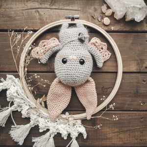 May include: A handmade crochet bat with gray body and pink wings and ears, set inside a wooden embroidery hoop. The bat has large black eyes and a small purple crystal. The background is a rustic wooden surface.