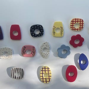 Könnte beinhalten: Eine Sammlung verschiedener kleiner, flacher, dekorativer Formen in Farben wie Rot, Grün, Violett, Gelb, Blau und Weiß. Diese Objekte weisen diverse Muster wie Punkte, Karos, Streifen und florale Motive auf, wobei viele eine mittige Aussparung besitzen.
