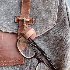 May include: A pair of eyeglasses with black frames and clear lenses is attached to a pink glasses holder. The holder is clipped to a brown leather strap on a gray canvas backpack.