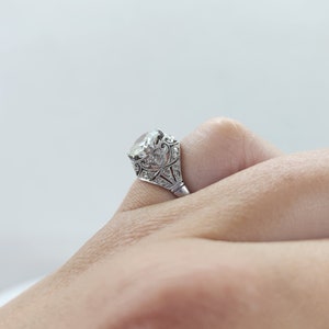 May include: A silver ring with a large, clear gemstone. The ring features intricate filigree and small accent stones around the main stone. The ring is set on a finger, showcasing its design and sparkle. A classic piece of jewelry.