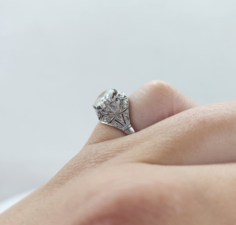 May include: A silver ring with a large, clear gemstone. The ring features intricate filigree and small accent stones around the main stone. The ring is set on a finger, showcasing its design and sparkle. A classic piece of jewelry.