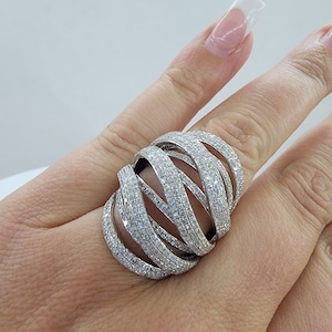 May include: A large, silver-toned ring with a woven design. The ring is encrusted with numerous small, clear stones, creating a sparkling effect. The ring's design features multiple bands that intertwine, forming a bold and eye-catching piece of jewelry.