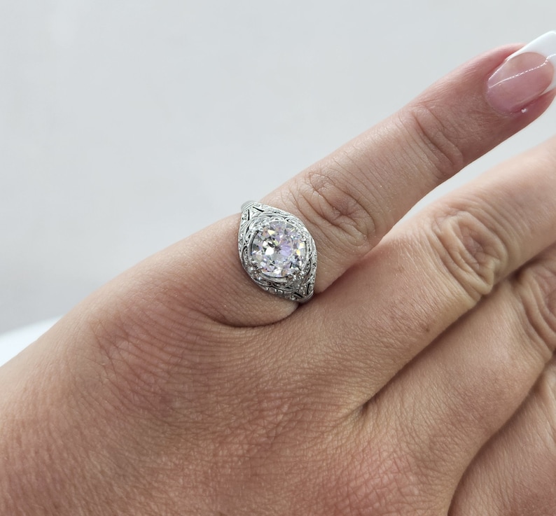 May include: A silver ring with a large, round, clear gemstone. The ring has an ornate filigree design around the gemstone and on the band. The ring is on a finger.