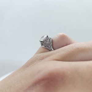 May include: Close-up of a silver ring with a large, clear gemstone. The ring features intricate filigree detailing around the stone and along the band. The ring is on a finger, showcasing the design and craftsmanship.