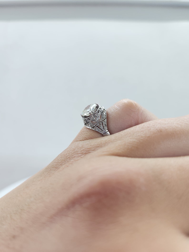 May include: Close-up of a silver ring with a large, clear gemstone. The ring features intricate filigree detailing around the stone and along the band. The ring is on a finger, showcasing the design and craftsmanship.