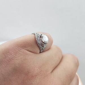 May include: A silver ring with an ornate filigree design. The ring features a large, round, clear gemstone as the centerpiece, surrounded by smaller accent stones. The ring is held by a hand against a white background.