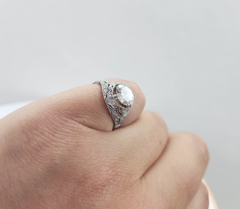 May include: A silver ring with an ornate filigree design. The ring features a large, round, clear gemstone as the centerpiece, surrounded by smaller accent stones. The ring is held by a hand against a white background.