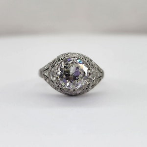 May include: A vintage-style engagement ring with a large, round-cut diamond as the centerpiece. The diamond is set in a detailed, filigree-style band, showcasing intricate metalwork. The ring's design is classic and elegant, with a focus on the brilliance of the diamond.
