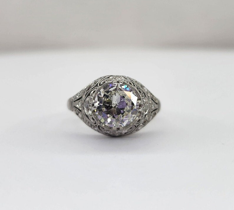 May include: A vintage-style engagement ring with a large, round-cut diamond as the centerpiece. The diamond is set in a detailed, filigree-style band, showcasing intricate metalwork. The ring's design is classic and elegant, with a focus on the brilliance of the diamond.