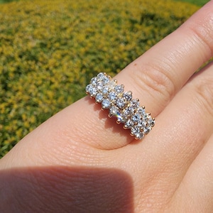 May include: A gold-toned ring with multiple clusters of clear, sparkling gemstones. The ring is set on a finger, with a blurred green background suggesting an outdoor setting. The gemstones catch the light, creating a brilliant display.