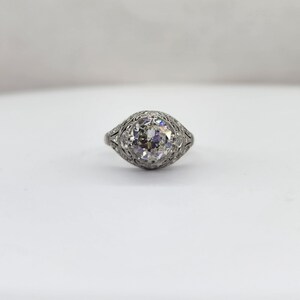 May include: A vintage-style ring with a large, round, clear diamond as the centerpiece. The ring's band and setting are intricately detailed with delicate filigree patterns. The ring is set against a plain white background.