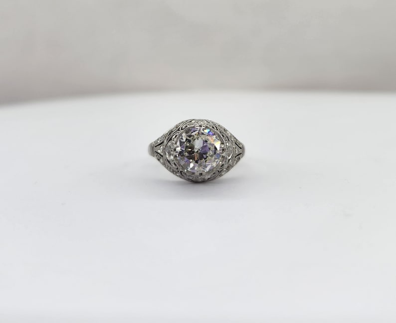 May include: A vintage-style ring with a large, round, clear diamond as the centerpiece. The ring's band and setting are intricately detailed with delicate filigree patterns. The ring is set against a plain white background.