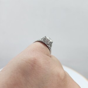 May include: A silver engagement ring with a large, clear, square-cut center stone. The ring features intricate filigree detailing and smaller accent stones along the band. The ring is shown on a finger.