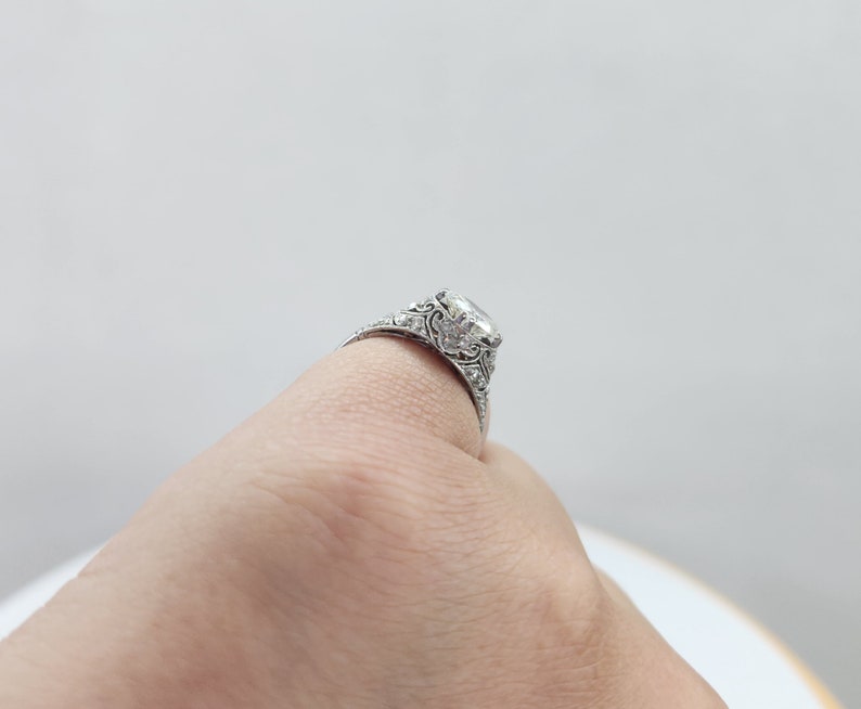 May include: A silver engagement ring with a large, clear, square-cut center stone. The ring features intricate filigree detailing and smaller accent stones along the band. The ring is shown on a finger.