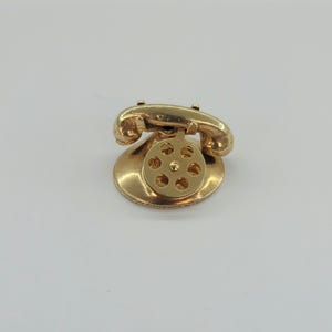 May include: A gold-colored vintage telephone charm. The rotary dial phone has a classic design with a handset, receiver, and a circular dial with holes. The charm is likely made of metal and has a shiny finish.