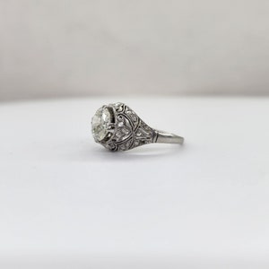 May include: A vintage-style engagement ring with a large, round, clear gemstone. The ring features intricate filigree and small accent stones set in a silver-toned metal. The design is ornate and detailed, showcasing a classic, elegant aesthetic.
