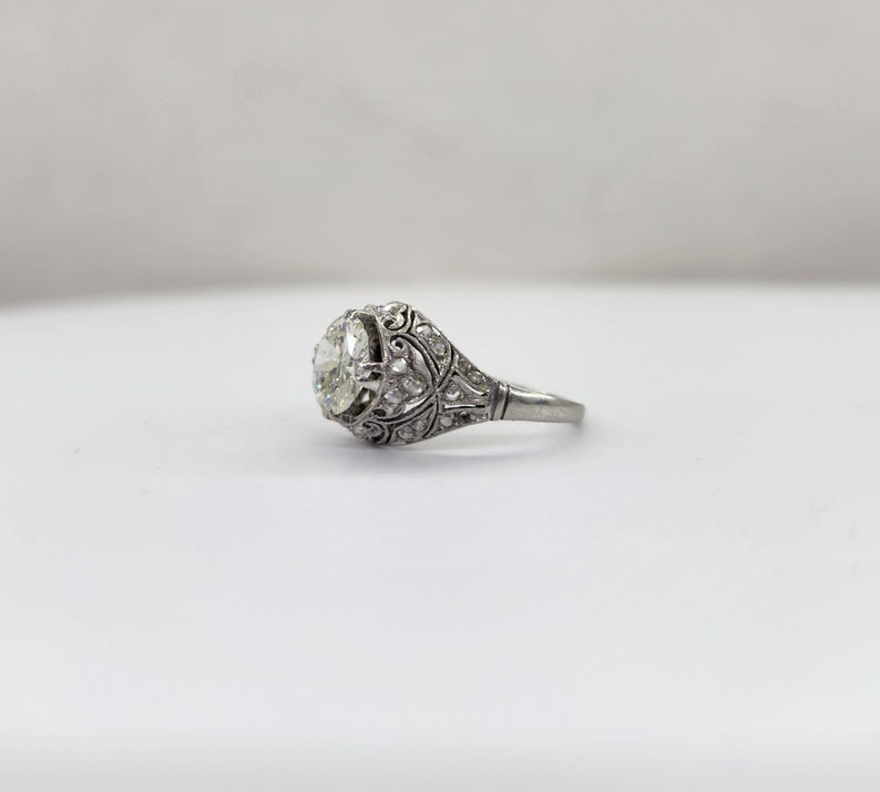 May include: A vintage-style engagement ring with a large, round, clear gemstone. The ring features intricate filigree and small accent stones set in a silver-toned metal. The design is ornate and detailed, showcasing a classic, elegant aesthetic.