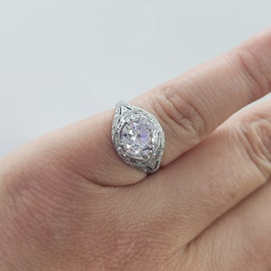 May include: A silver engagement ring with a large, round, clear gemstone. The ring features intricate filigree detailing on the band and around the gemstone. The ring is shown on a finger against a white background.