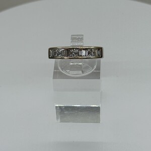 May include: A gold-toned ring with a row of rectangular-cut gemstones. The ring is displayed on a clear acrylic stand against a white background. The gemstones are set in a channel setting.