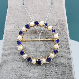 May include: A gold circle brooch with alternating blue and white stones. The brooch is on a gray background.