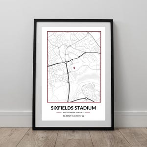 May include: Framed print showing a detailed map of the Sixfields Stadium area. The map is in black and white with a red marker indicating the stadium's location. The text "SIXFIELDS STADIUM" is displayed at the bottom, with coordinates.