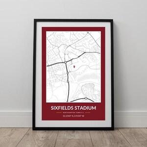 May include: Framed print showing a black and white map with a red border and a red banner. The banner reads "SIXFIELDS STADIUM" and includes coordinates. The map details roads and buildings, with a marker indicating a location.